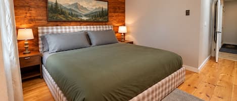 A bedroom with a wooden headboard, a bed with a green bedspread, two bedside lamps, and a painting of a mountain landscape.