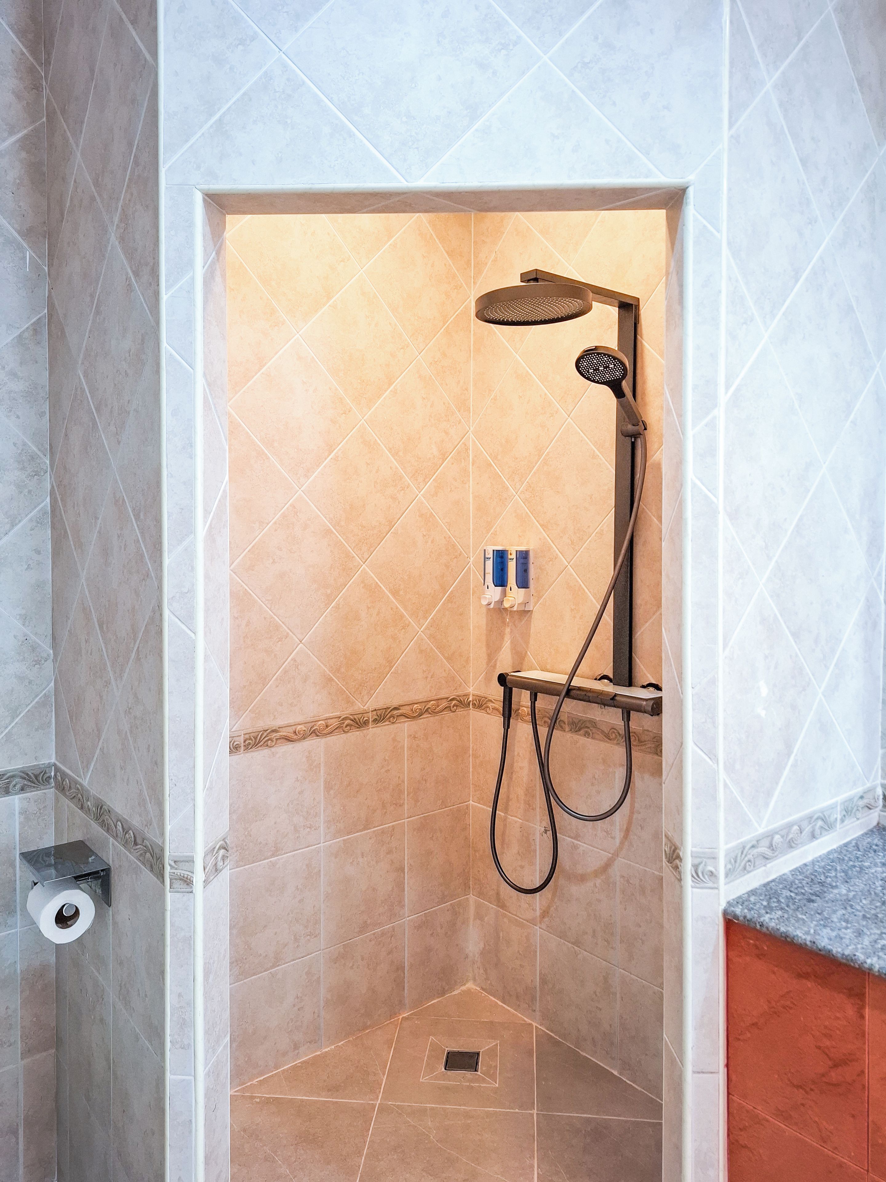 A shower area with a handheld showerhead, a wall-mounted control panel, and a corner tile design.