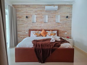 A hotel room with a brick-patterned wall, a bed with white bedding and brown headboard, and a nightstand with a glass of red wine. - Beachside 4BR Private Pool Villa  (Muang Pattaya)
