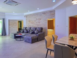 A modern living room with a sectional sofa, a round coffee table, and a dining area with a table and chairs. - Beachside 4BR Private Pool Villa  (Muang Pattaya)