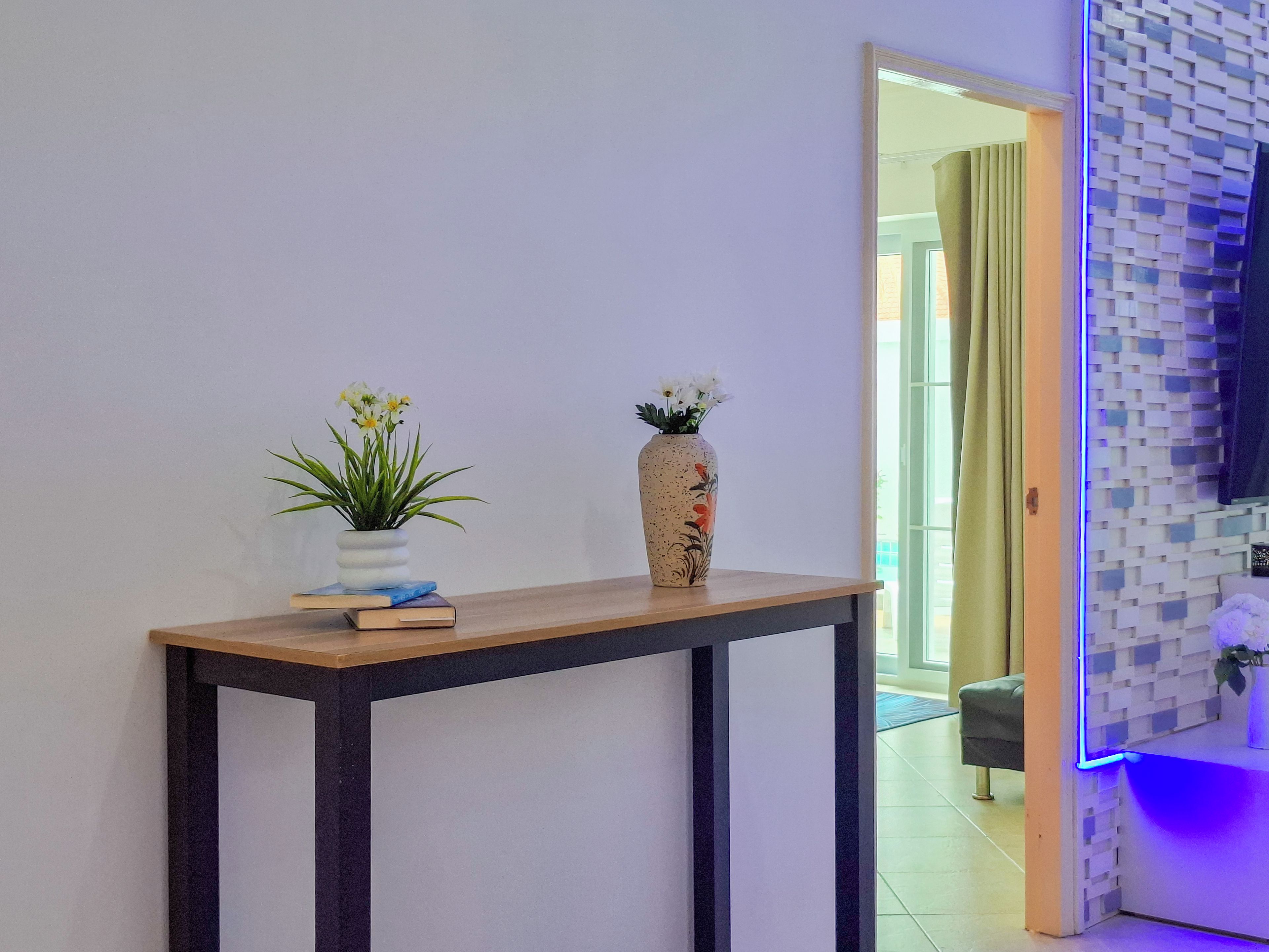 A hallway with a wooden console table, two vases with flowers, and a mirror reflecting a room with a patterned wall and a blue-lit corner.