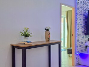 Ein Flur mit einem Holzkonsolentisch, zwei Vasen mit Blumen und einem Spiegel, der einen Raum mit einer gemusterten Wand und einer blau beleuchteten Ecke reflektiert.
