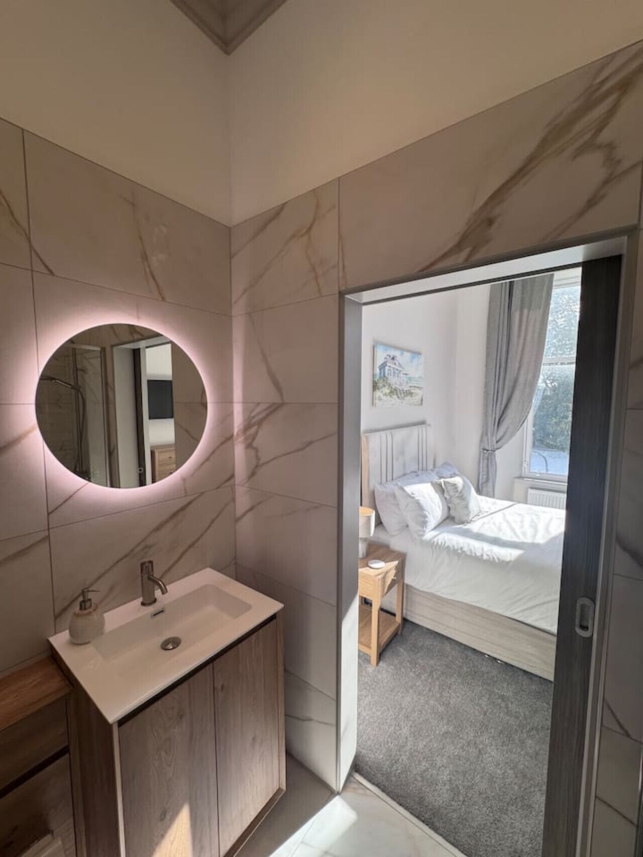A modern bathroom with a marble vanity, a round mirror, and a bed visible through an open door.