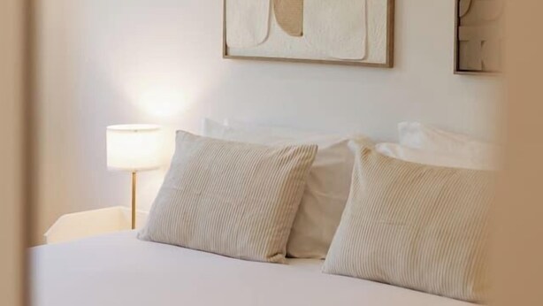 Quarto de hotel com cama, almofadas, mesa de cabeceira e uma obra de arte abstrata pendurada na parede.