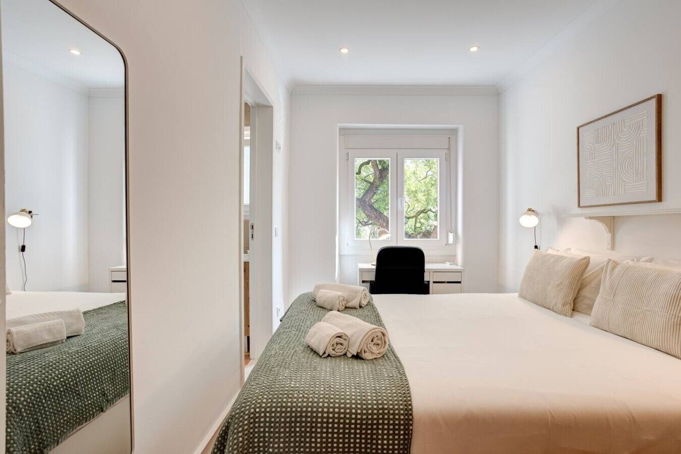 Een hotelkamer met een groot bed, een bureau, een stoel, een raam met uitzicht op bomen, en een spiegel.