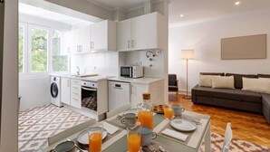 Cozinha moderna com armários brancos, sala de jantar com mesa posta para o café da manhã, e sala de estar com sofá e abajur.