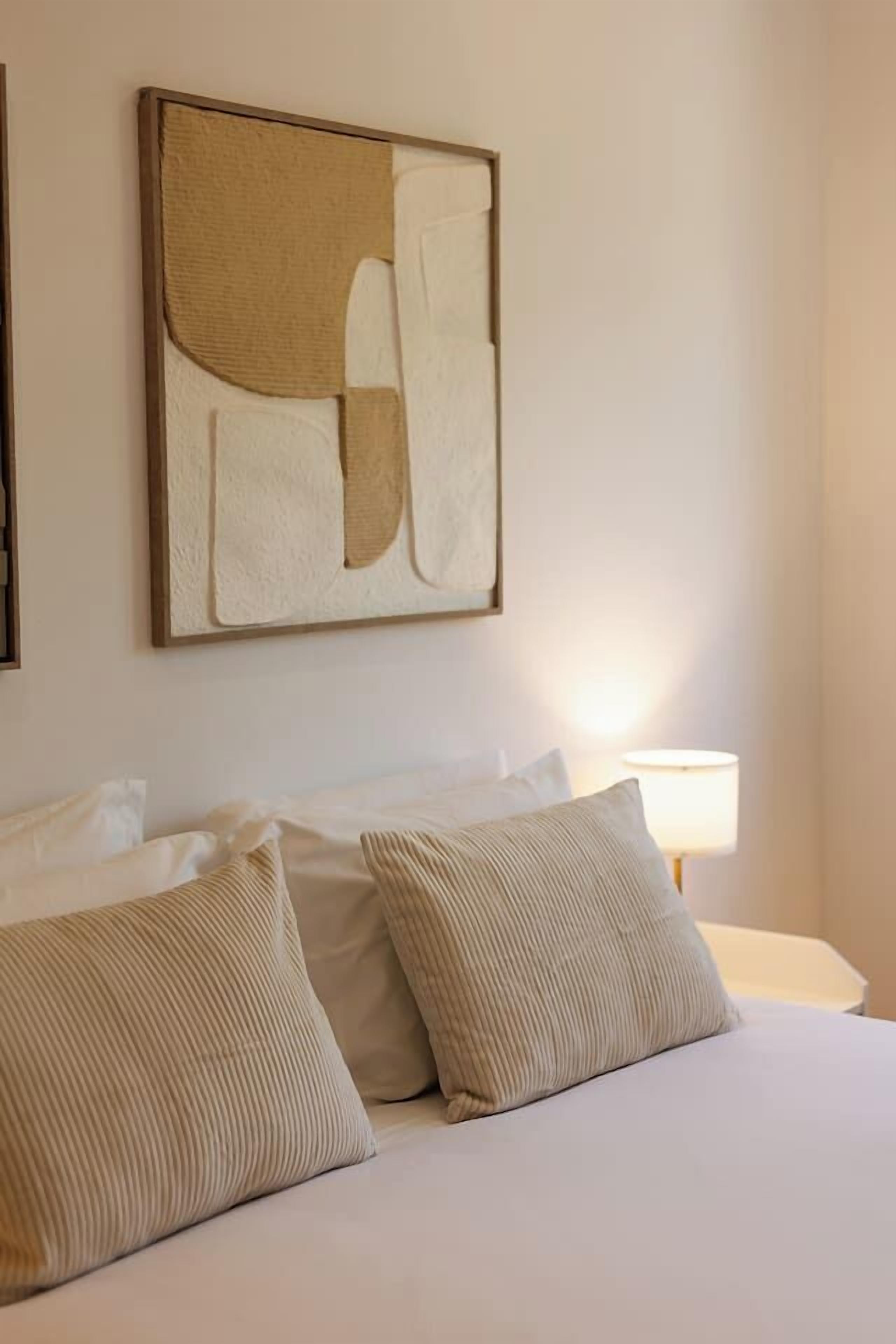 Ein Hotelzimmer mit einem Bett, Kissen, einem Wandbild im abstrakten Stil und einer Nachttischlampe.