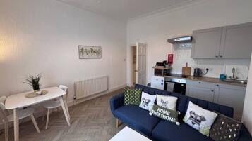 A compact living space with a dining area, a sofa with decorative pillows, a small white table, and a kitchenette in the background.