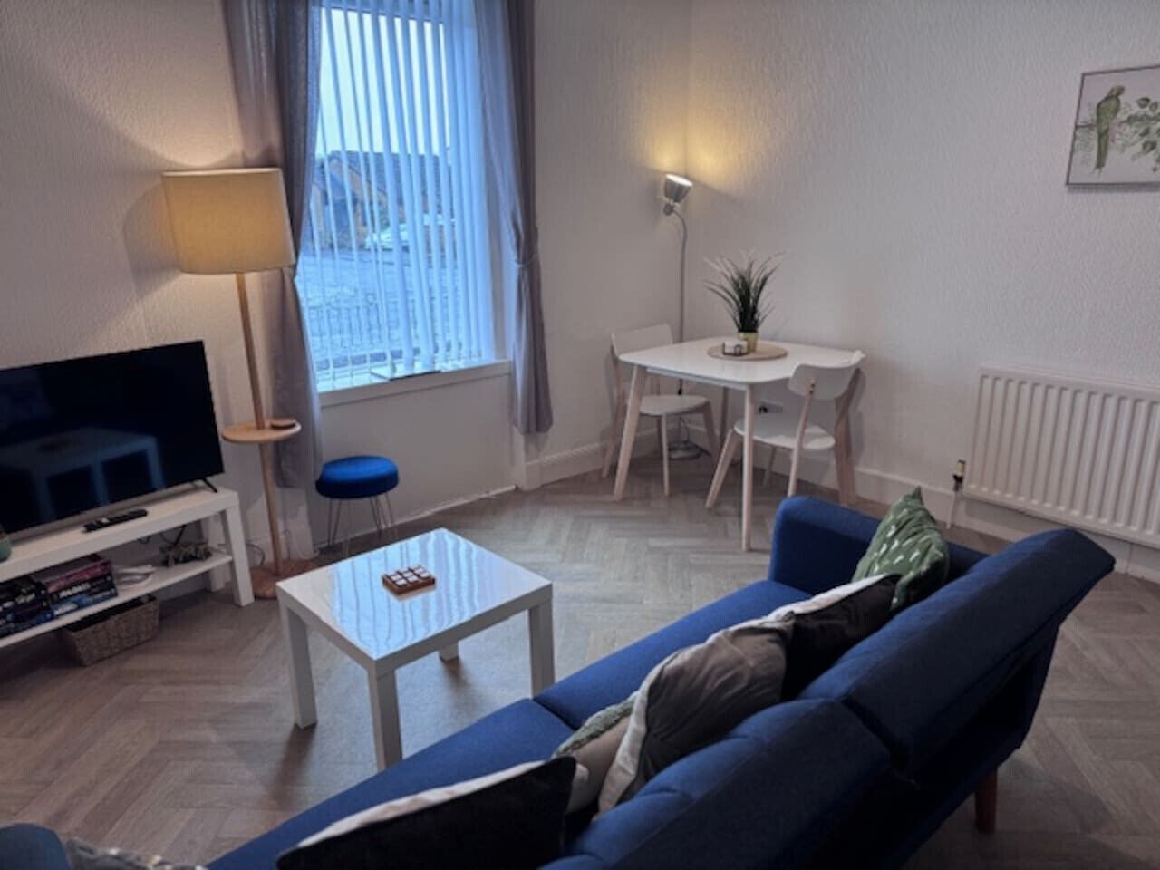 A living room with a TV, a sofa, a coffee table, a dining table with chairs, and a window with blinds.