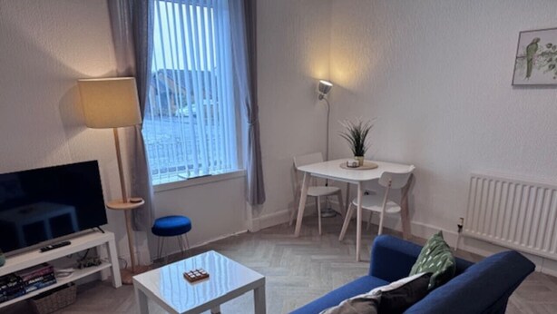 En stue med et fjernsyn, en sofa, et sofabord, et spisebord med stole og et vindue med persienner.