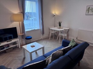 A living room with a TV, a sofa, a coffee table, a dining table with chairs, and a window with blinds. - The wee Balloch But & Ben - Macaulay Letting (Balloch)