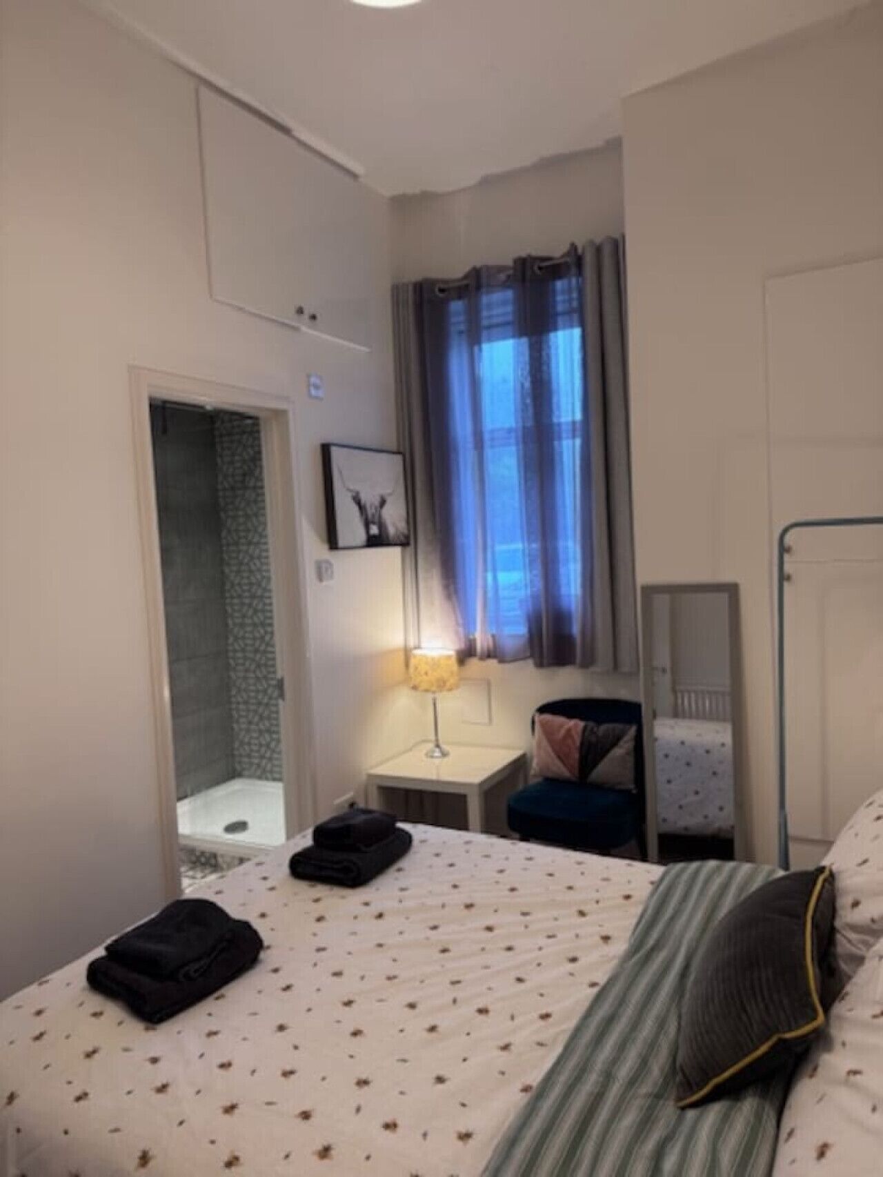 Et hotelværelse med en seng, et natbord, en lampe, et brusebadeværelse og et vindue med gardiner.