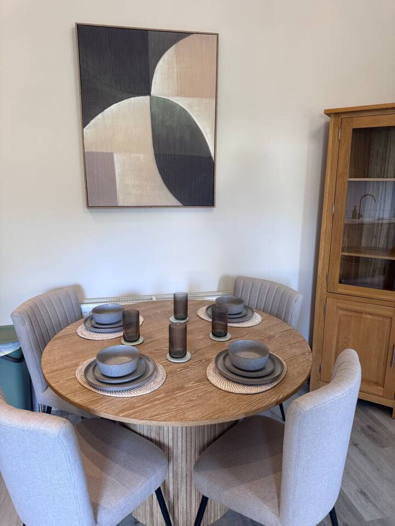 Una mesa de comedor redonda con seis sillas, un aparador de madera y un cuadro abstracto colgado en la pared.