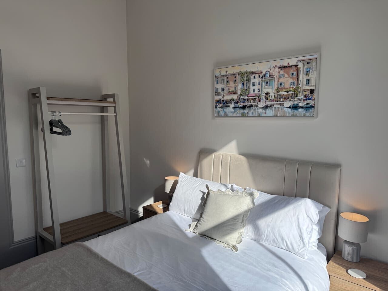 Un dormitorio con cama, mesita de noche, lámpara, una obra de arte en la pared y un perchero.