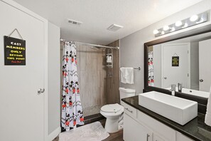 Une salle de bain avec un rideau de douche orné de personnages de dessins animés, des toilettes, un lavabo avec un miroir et une pancarte sur la porte.