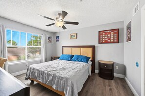 Une chambre à coucher comprenant un lit, une table de chevet, un ventilateur de plafond, une fenêtre avec des stores et un mur orné de tableaux.