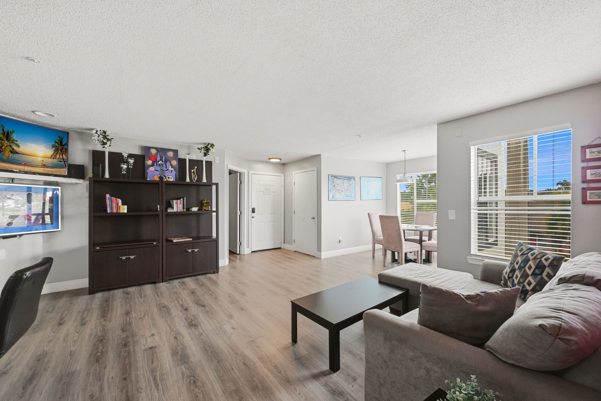 A modern living room with a dark wood floor, a sofa, a coffee table, and a bookshelf. There is a dining area with a table and chairs, and a window with blinds.