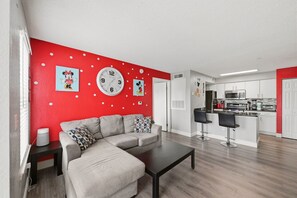 Ein modernes Wohnzimmer mit einer Eckcouch, einem Couchtisch und einer Küchenzeile inklusive Mikrowelle, Herd und Barhocker.