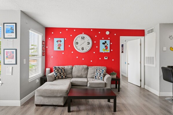 Un salon avec un mur d’accent rouge, un canapé d’angle gris, une horloge murale ronde et des tableaux encadrés.