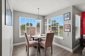 Un coin repas avec une table ronde et quatre chaises, de grandes fenêtres équipées de stores et des œuvres d’art accrochées au mur.