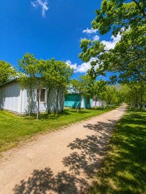 Una hilera de cabañas blancas con un sendero de tierra entre ellas, rodeadas de árboles verdes y un cielo azul despejado.