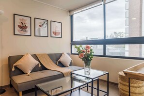 Een moderne woonkamer met een bank, een glazen salontafel en een raam met uitzicht op gebouwen.