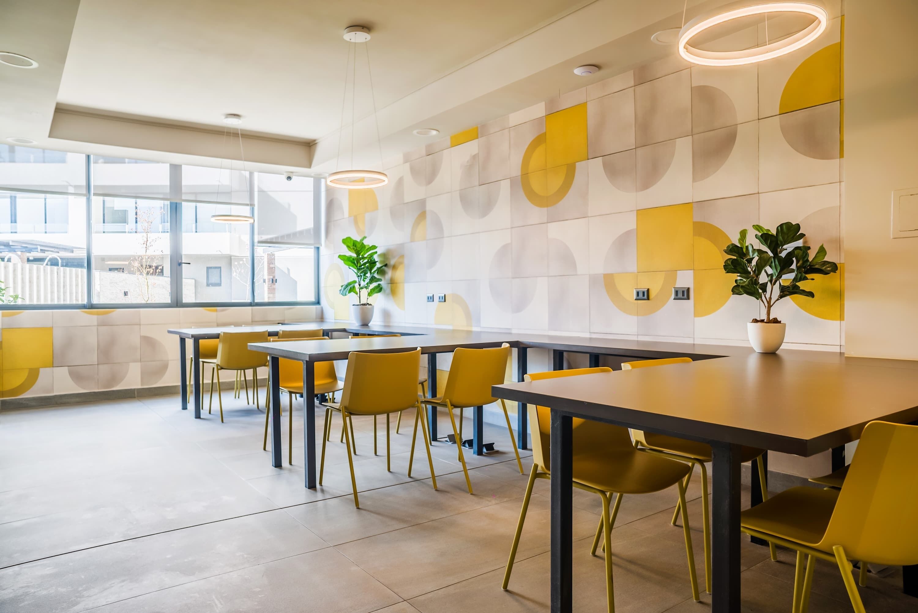 Un'area pranzo moderna con un motivo geometrico sulla parete, sedie gialle e lampade a sospensione.
