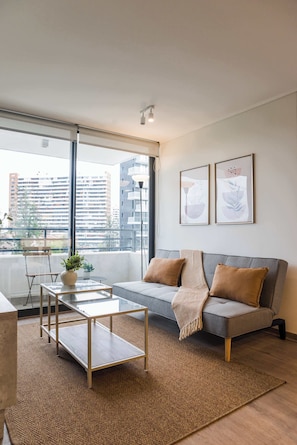 A modern living room with a grey sofa, a glass coffee table, and a view of buildings through large windows. - Beautiful 2BR Apt in Las Condes with Pool & Views (Las Condes)