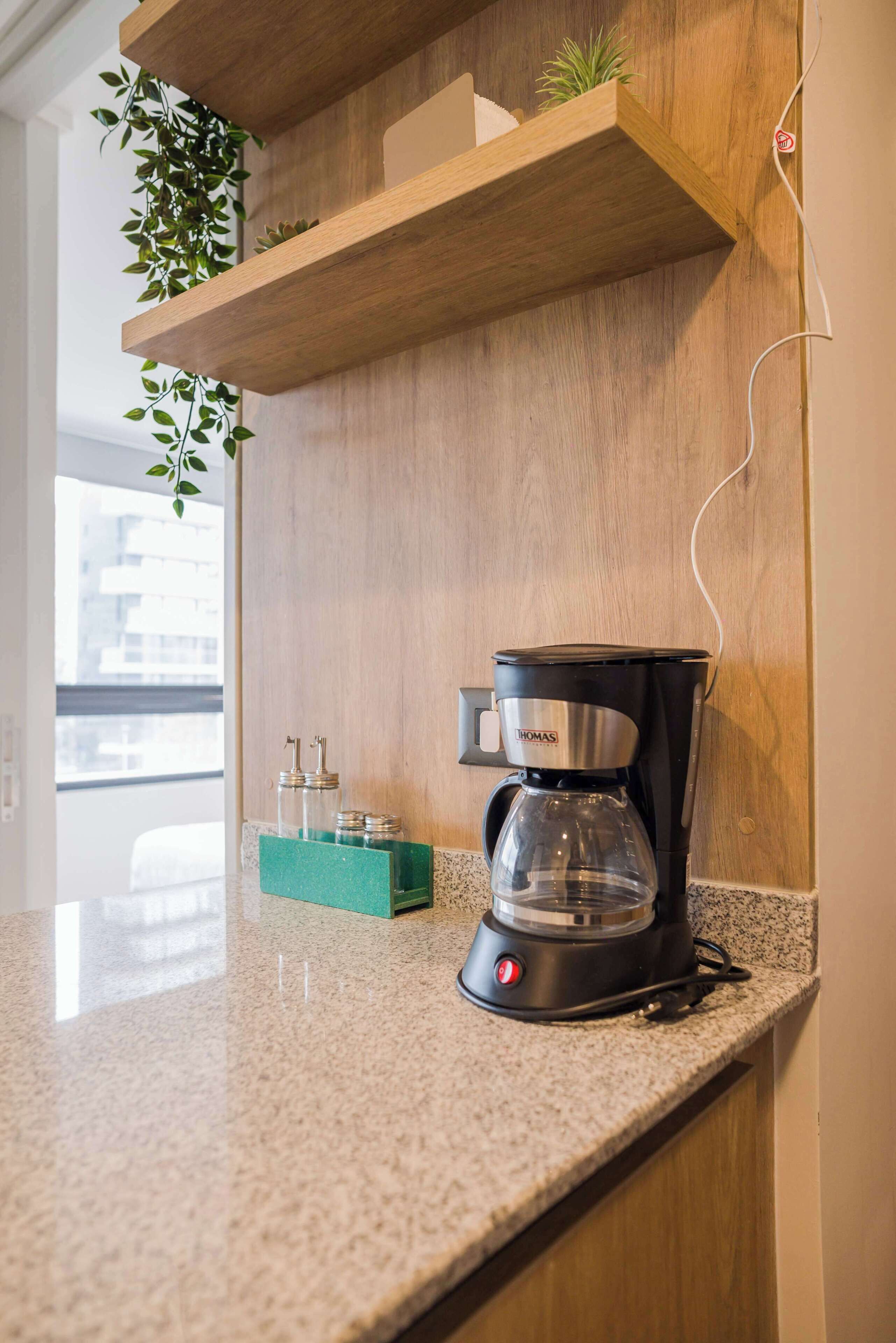 A kitchen with a coffee maker on the counter, a plant on the shelf, and a window with a view.