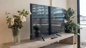 A flat-screen TV mounted on a wooden console with a remote on it. There are two potted plants on either side of the TV. - Beautiful 2BR Apt in Las Condes with Pool & Views (Las Condes)