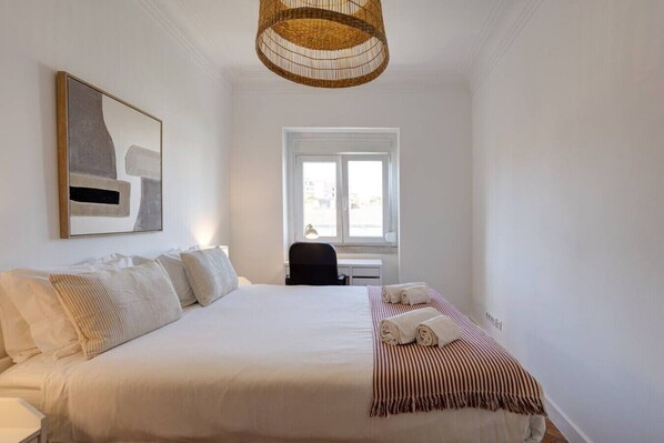 A hotel room with a large bed, a desk, a chair, a window, and a hanging light fixture. - Bright, Custom-Built Apartment with Air Conditioning in Areeiro (Lisbon)