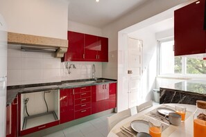 A modern kitchen with red cabinets, white tiles, and a dining area with a table set for two. - Bright, Custom-Built Apartment with Air Conditioning in Areeiro (Lisbon)