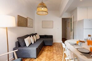 A modern living room with a grey sectional sofa, a herringbone wood floor, and a dining area with a white table set for breakfast. - Bright, Custom-Built Apartment with Air Conditioning in Areeiro (Lisbon)