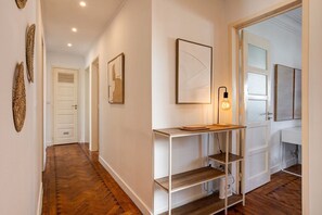 A hallway with a wooden floor, white walls, and a white door at the end. There are framed artworks on the walls and a table with a lamp. - Bright, Custom-Built Apartment with Air Conditioning in Areeiro (Lisbon)