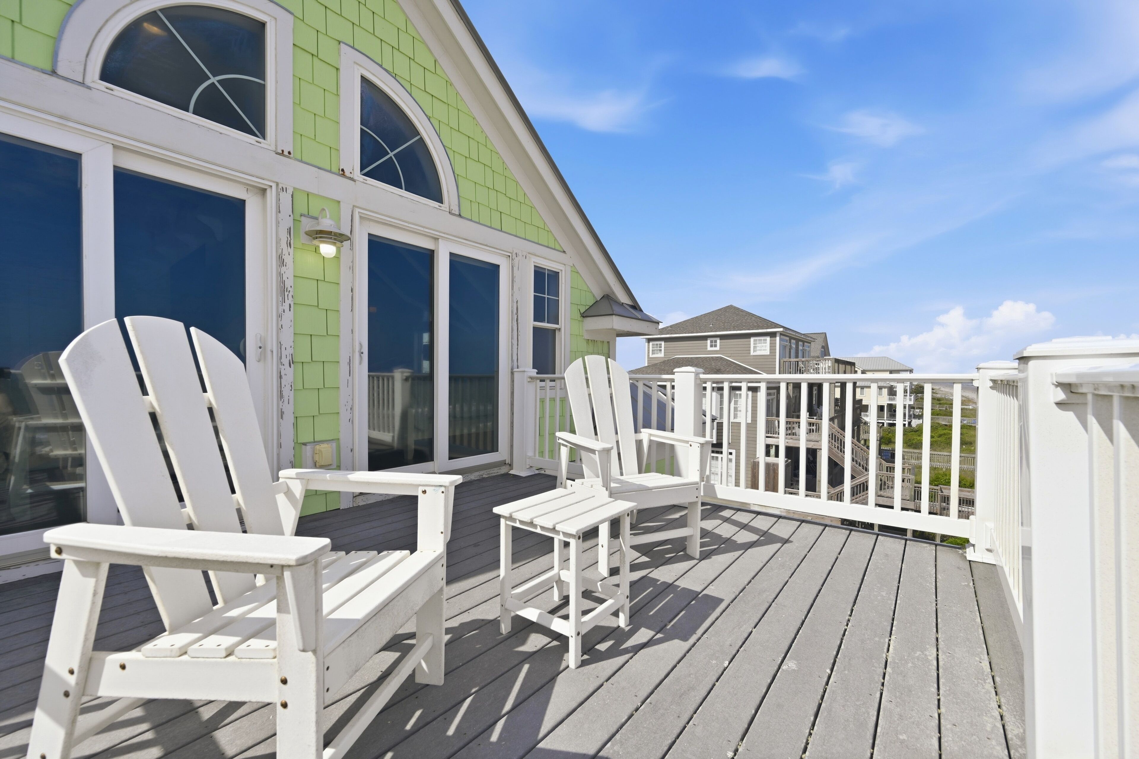 Eine Holzterrasse mit weißen Adirondack-Stühlen und einem Gebäude mit großen Fenstern in Grün.