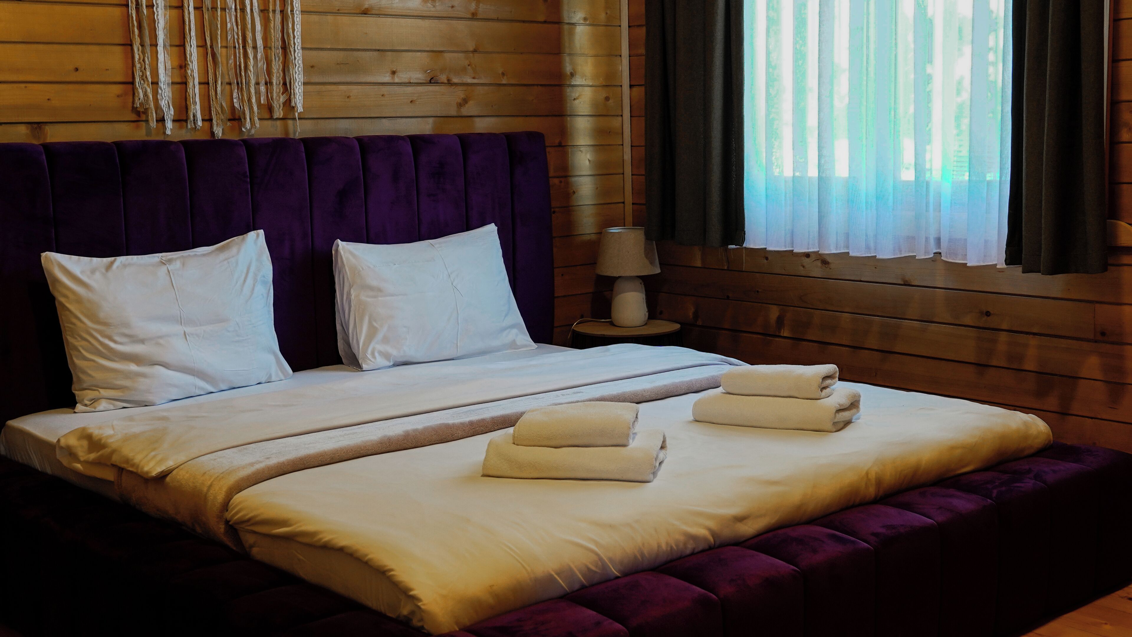 Una cama doble con sábanas blancas, un cabecero morado y dos almohadas. Hay dos toallas dobladas sobre la cama.