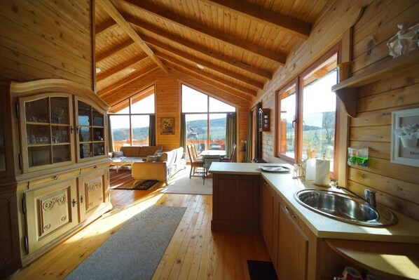 A wooden cabin interior with a kitchen, dining area, and a view of the outdoors.