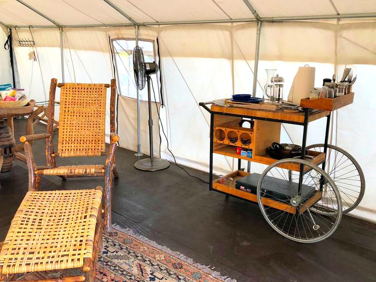 Ein Zeltinnenraum mit einem Korbstuhl, einem kleinen Tisch mit einem Ventilator und einem Wagen mit Regalen.