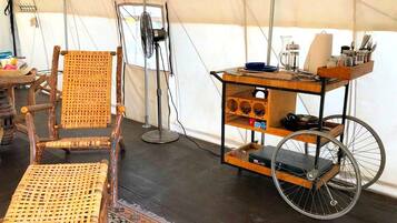 Ein Zeltinnenraum mit einem Korbstuhl, einem kleinen Tisch mit einem Ventilator und einem Wagen mit Regalen.