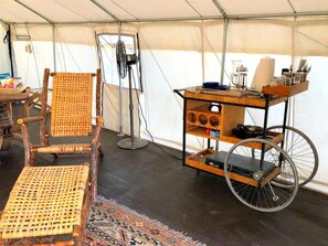 Ein Zeltinnenraum mit einem Korbstuhl, einem kleinen Tisch mit einem Ventilator und einem Wagen mit Regalen.
