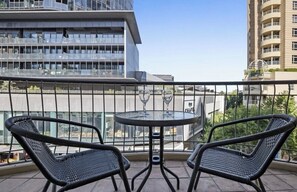 Un balcó amb dues cadires de vimetla i una taula petita, amb vista a un edifici modern de diversos pisos.