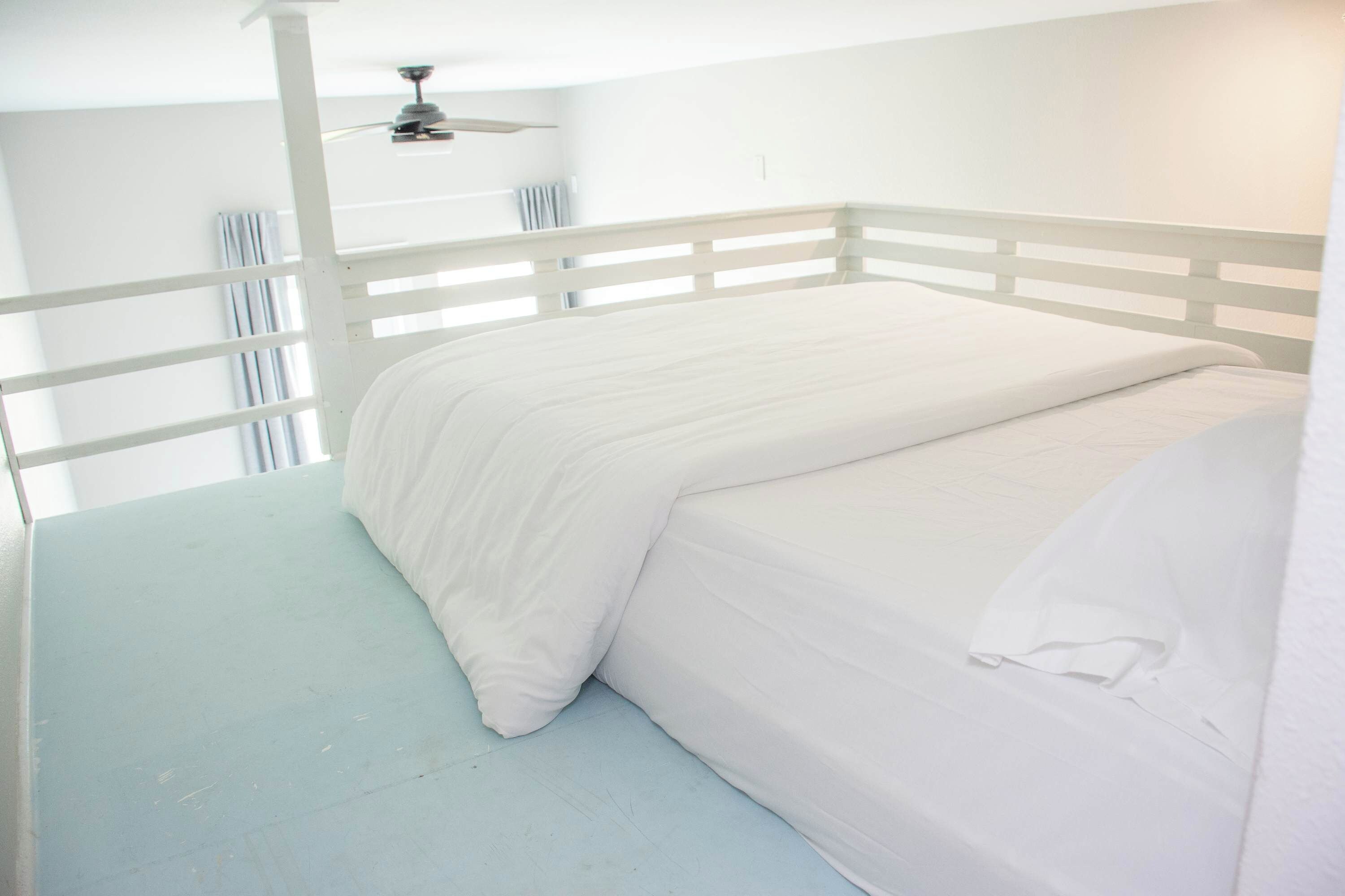 A single-bed room with a white ceiling fan, white walls, and a light blue floor.