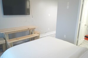 A bedroom with a bed, a wooden bench, a television mounted on the wall, and a door leading to another room.