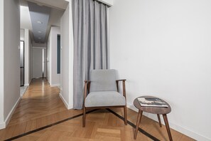 A hallway with a grey upholstered chair, a small round table with a magazine, and a grey curtain. - Lumina 3room Apart with Balkony & Center (Warszawa)
