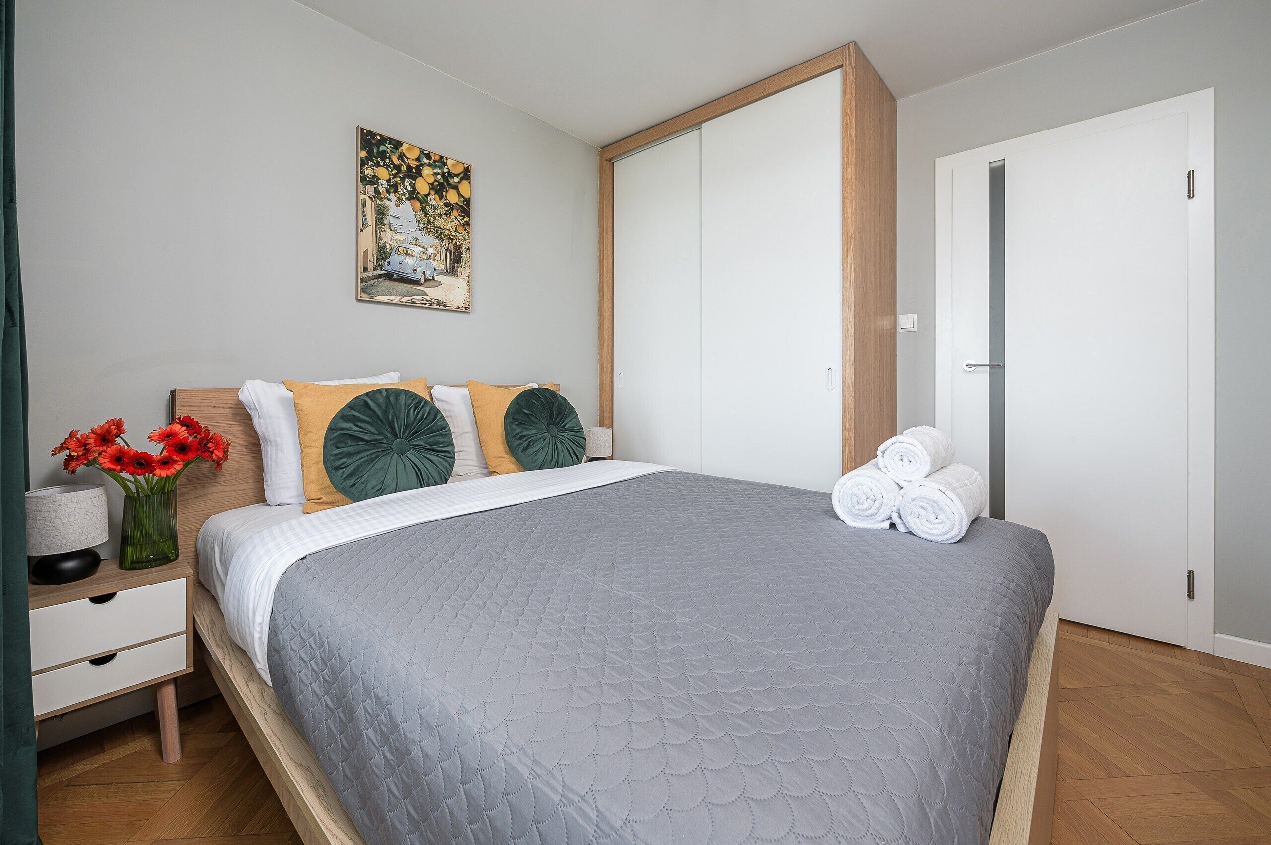 A neatly arranged bedroom with a bed, pillows, a bedside table with a lamp, a vase with flowers, and a painting on the wall.