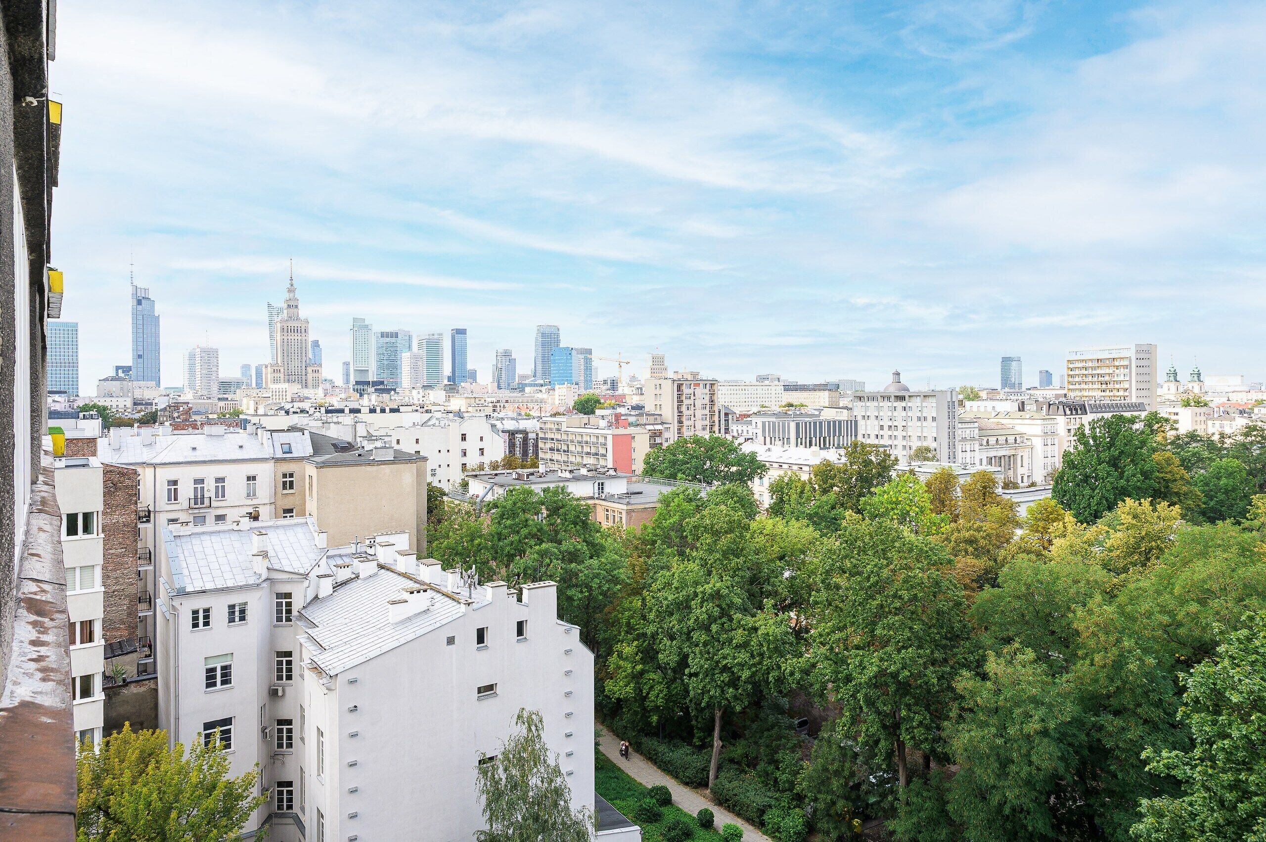 A cityscape with a mix of modern and older buildings, a clear sky, and a tree-lined foreground.
