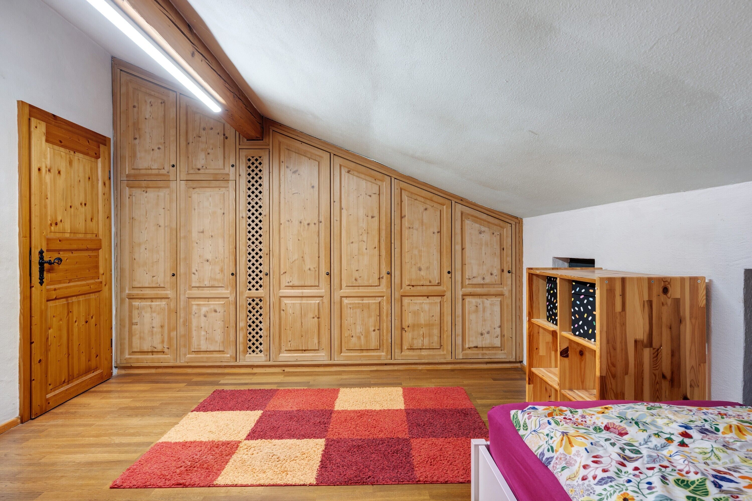 A bedroom with a wooden wardrobe, a checkered rug, and a bed with a floral bedspread.