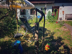 Un espacio al aire libre rústico con un parasol de paja, una silla plegable y una herramienta de metal apoyada en un árbol.
