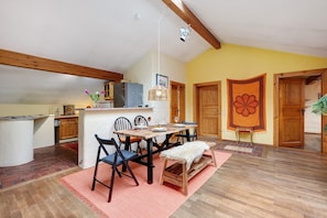 A dining area with a wooden table, chairs, and a bench. There is a kitchen area with a refrigerator and a hanging light fixture.