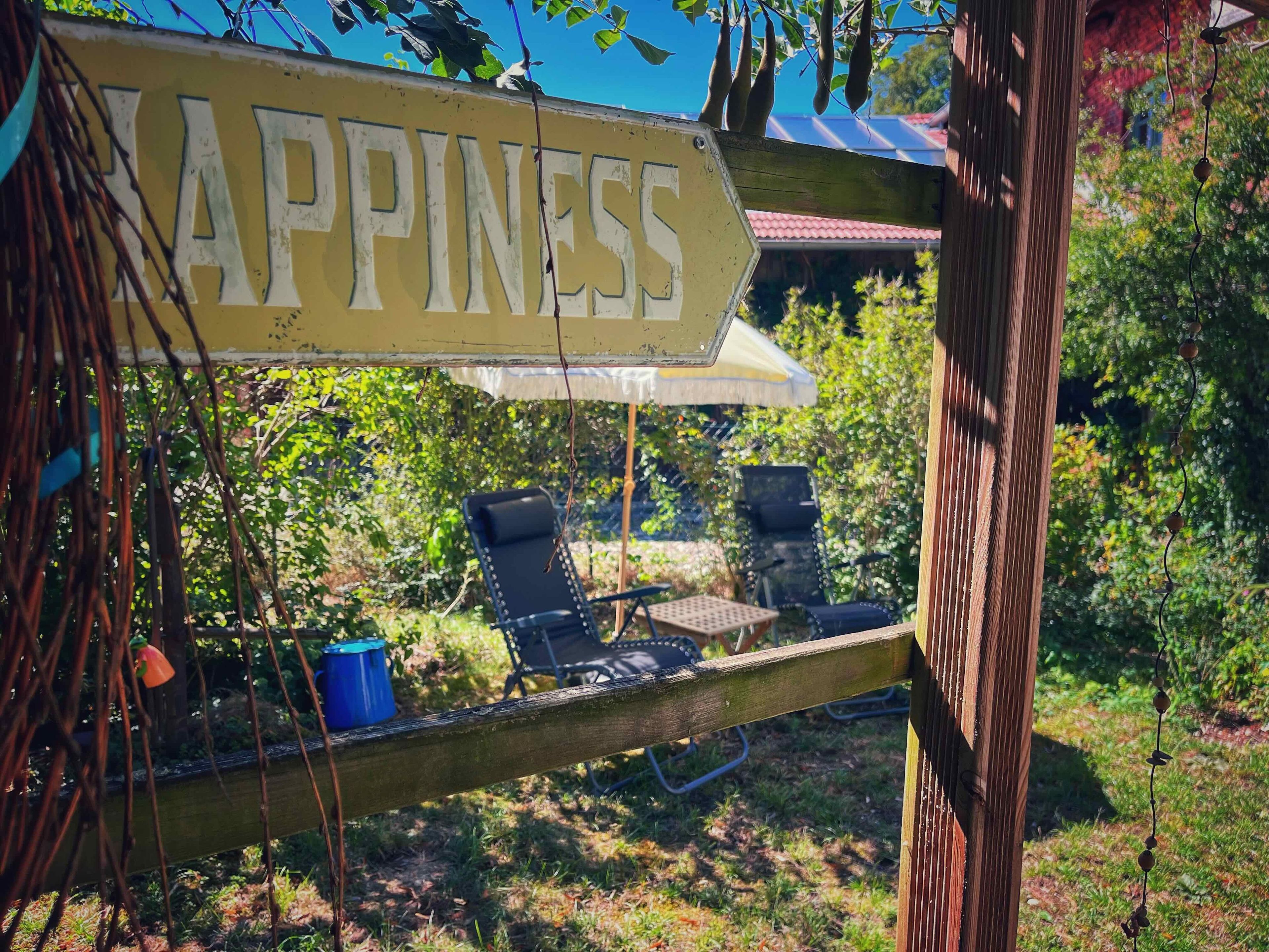 Une terrasse en bois avec une pancarte sur laquelle on peut lire « BONHEUR », une table, deux chaises et un parasol suspendu.